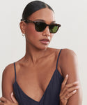 A woman with dark, pulled-back hair wears Broadway Sunglasses by Garrett Leight, a navy spaghetti-strap top, and gold hoop earrings, crossing her arms and gazing to the side against a plain light background.
