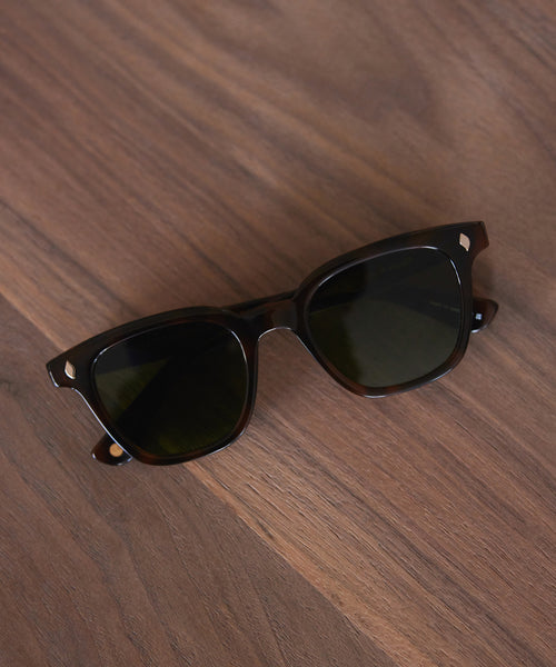 The Broadway Sunglasses by Garrett Leight, featuring black frames and dark UV-protective lenses, rest on a brown wooden surface.