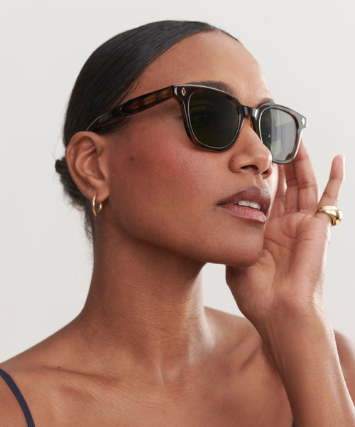 A woman with smooth, dark skin and slicked-back hair wears Broadway Sunglasses, offering UV protection. Gold hoop earrings and a gold ring add elegance as she gently touches her face and gazes confidently to the side.