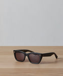 A pair of Webster Sunglasses with black rectangular acetate frames and dark lenses rests on a light wooden surface against a plain, light gray background.