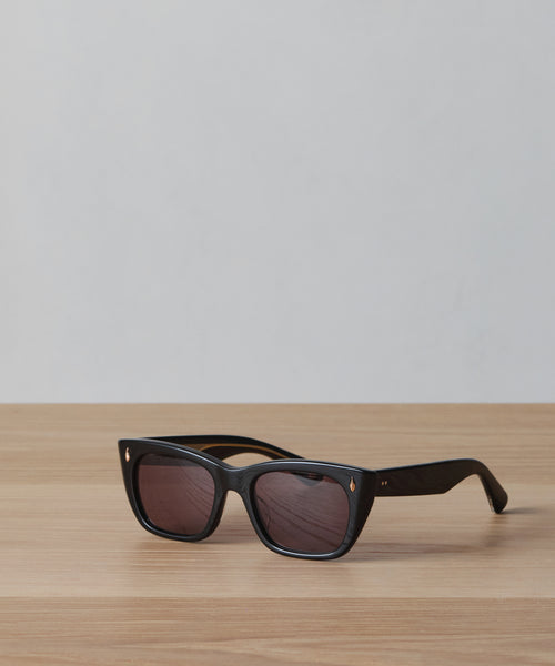 A pair of Webster Sunglasses with black rectangular acetate frames and dark lenses rests on a light wooden surface against a plain, light gray background.