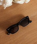 A pair of Webster Sunglasses with black acetate frames and dark lenses rests on a wooden surface. White flowers in the top left add a fresh accent to these stylish eyewear frames.
