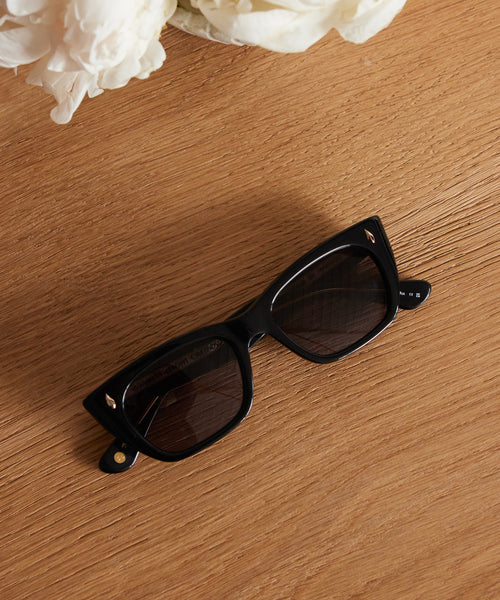 A pair of Webster Sunglasses with black acetate frames and dark lenses rests on a wooden surface. White flowers in the top left add a fresh accent to these stylish eyewear frames.