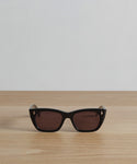 The Webster Sunglasses, in sleek black rectangular acetate, feature dark lenses and subtle gold accents, elegantly shown on a light wooden surface with a plain white background.