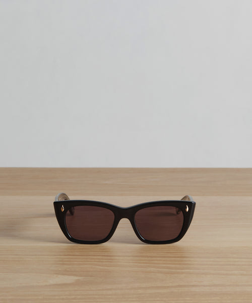 The Webster Sunglasses, in sleek black rectangular acetate, feature dark lenses and subtle gold accents, elegantly shown on a light wooden surface with a plain white background.