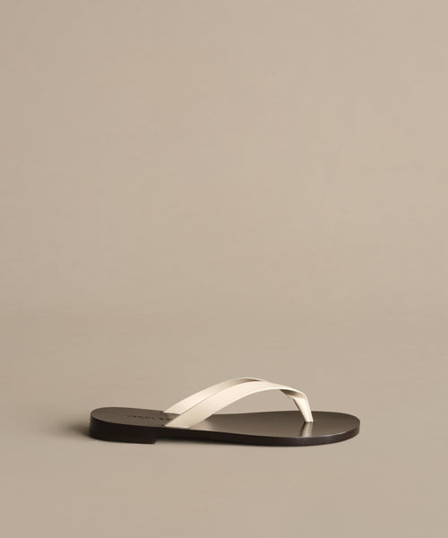 The Gia Sandal features white crisscross straps and a flat black sole, showcased on a light taupe background.