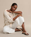A woman sits on the floor against a beige background, wearing the classic Gia Sandal paired with a cream and beige striped sweater and white wide-leg pants. She rests her arm on her knee, gazing at the camera with a gentle smile.