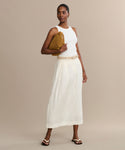 A woman stands against a beige background wearing a white sleeveless top, cream long skirt, and the Gia Sandal while holding a brown clutch and gazing to the side with a neutral expression.