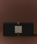 A rectangular black box with a gold vertical band and a white label reading Glass Candle Gift Set sits against a plain brown background, presenting an elegant candle gift set perfect for fans of scented candles.