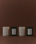 The Glass Candle Gift Set displays four minimalist candles (two white, two black) in a row on a brown background, each labeled with a unique scent: Ash, Aspen, Cedar, and Sun Valley.