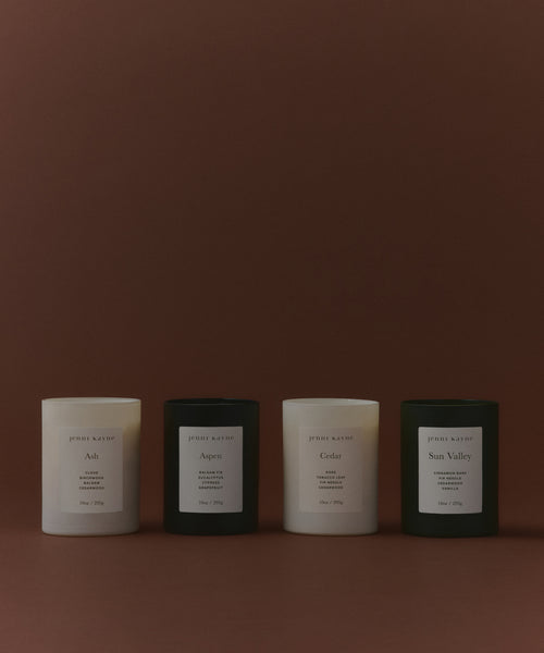 The Glass Candle Gift Set displays four minimalist candles (two white, two black) in a row on a brown background, each labeled with a unique scent: Ash, Aspen, Cedar, and Sun Valley.