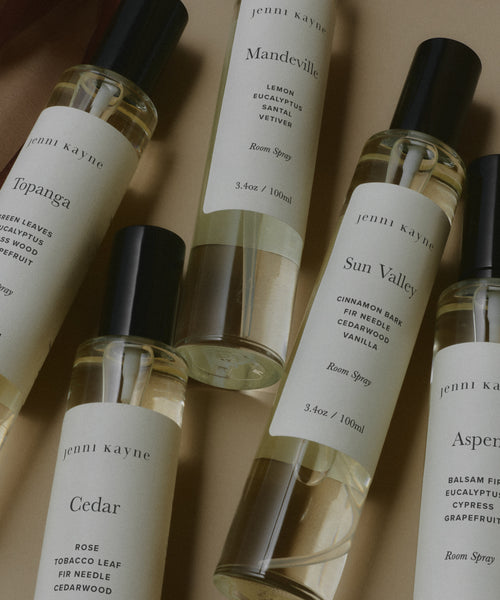 Five bottles from the Room Spray Sample Scent Set are arranged on a neutral surface, featuring Topanga, Cedar, Mandeville, Sun Valley, and Aspen—each labeled with its signature fragrance notes.