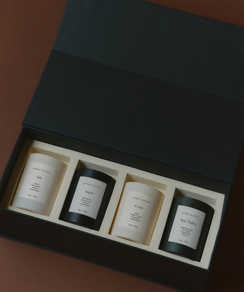 The Glass Candle Gift Set includes four scented candles in black and white jars labeled Ash, Aspen, Cedar, and Sun Valley, attractively arranged on a brown surface.