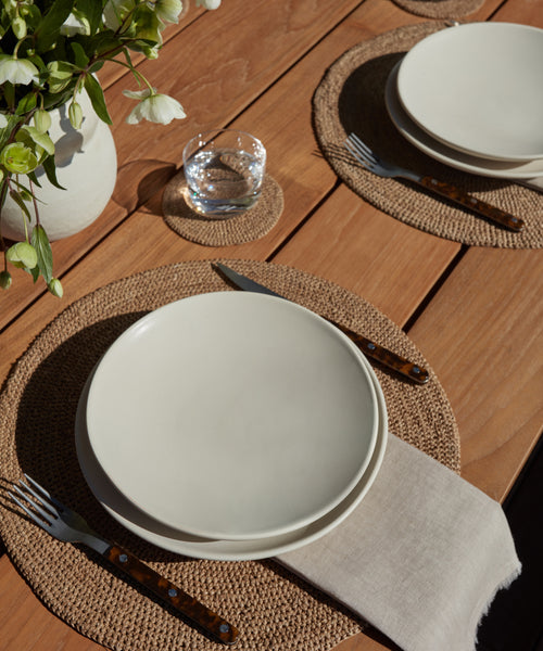 A wooden table set for two with white plates, woven placemats, brown-handled cutlery, linen napkins, and the Universal Low Glass Set of 4. Sunlight creates soft shadows beside a vase of white flowers.