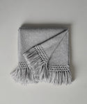 The Glenbrook Throw, in light gray with fringed edges and long tassels, is neatly folded on a white surface. One corner is turned over, showing off its textured woven pattern.