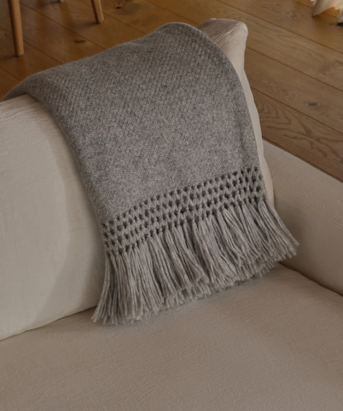 The Glenbrook Throw, a gray fringed woven blanket, is draped over the arm of a light beige upholstered sofa, with wooden flooring and chair legs visible in the background.