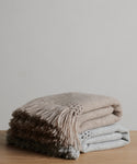 Two folded Glenbrook Throw blankets—one beige, one light gray—stacked on a wooden surface against a plain background. Each is a soft, woven throw designed for comfort and warmth, finished with fringe detailing.