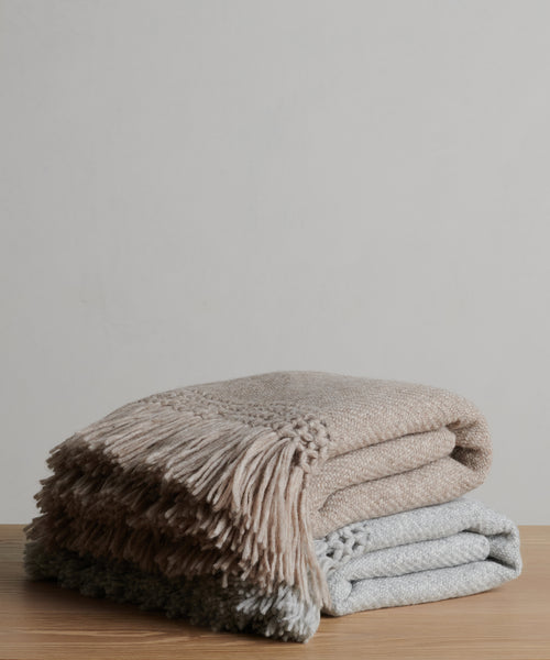 Two folded Glenbrook Throw blankets—one beige, one light gray—stacked on a wooden surface against a plain background. Each is a soft, woven throw designed for comfort and warmth, finished with fringe detailing.