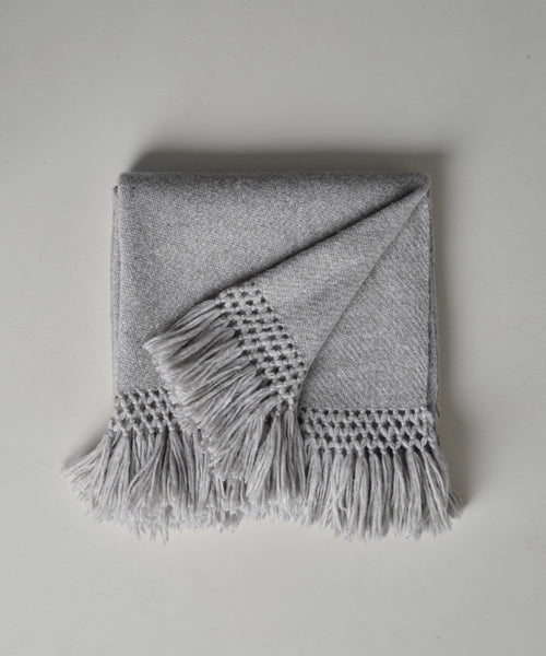 The Glenbrook Throw, in light gray with fringed edges and long tassels, is neatly folded on a white surface. One corner is turned over, showing off its textured woven pattern.