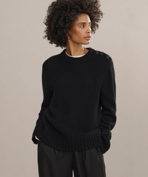 Wearing the Grace Crewneck, a black cashmere sweater with shoulder button details, and loose black pants, a woman with curly hair stands against a beige background, hands in her pockets—an effortless luxury knitwear look.