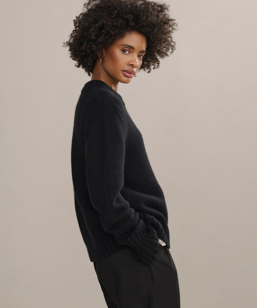 A woman with curly hair stands sideways against a beige background wearing the Grace Crewneck in black and matching pants, looking toward the camera with a neutral expression—showcasing understated luxury knitwear.