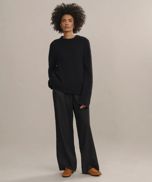 A woman with curly hair stands against a plain background, wearing the Grace Crewneck in black, wide-leg black pants, and brown shoes. Her left hand is in her pocket as she looks straight ahead with a neutral expression.