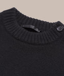 Close-up of the Grace Crewneck, a black cashmere sweater with a ribbed crew neckline and a single black shoulder button. The partially visible label highlights its luxury knitwear quality, set against a solid beige background.