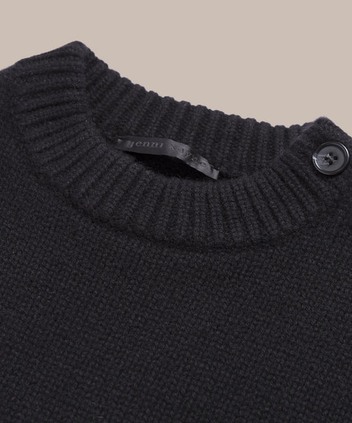 Close-up of the Grace Crewneck, a black cashmere sweater with a ribbed crew neckline and a single black shoulder button. The partially visible label highlights its luxury knitwear quality, set against a solid beige background.