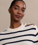 A person with short hair wears the Grace Crewneck, a luxury navy striped knit sweater with shoulder buttons, and small hoop earrings, posing against a plain beige background.