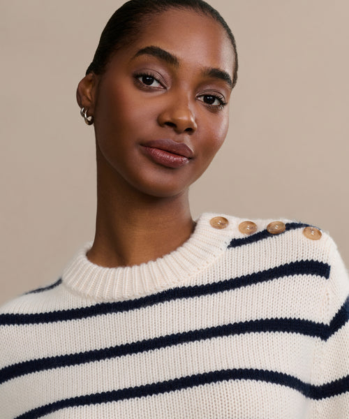 A person with short hair wears the Grace Crewneck, a luxury navy striped knit sweater with shoulder buttons, and small hoop earrings, posing against a plain beige background.