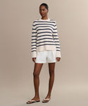 A woman wearing the Grace Crewneck in navy stripe, white shorts, and black flip-flops stands against a plain beige background.