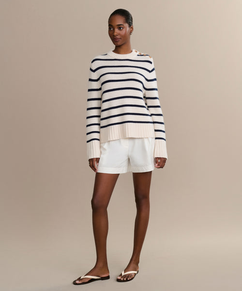 A woman wearing the Grace Crewneck in navy stripe, white shorts, and black flip-flops stands against a plain beige background.