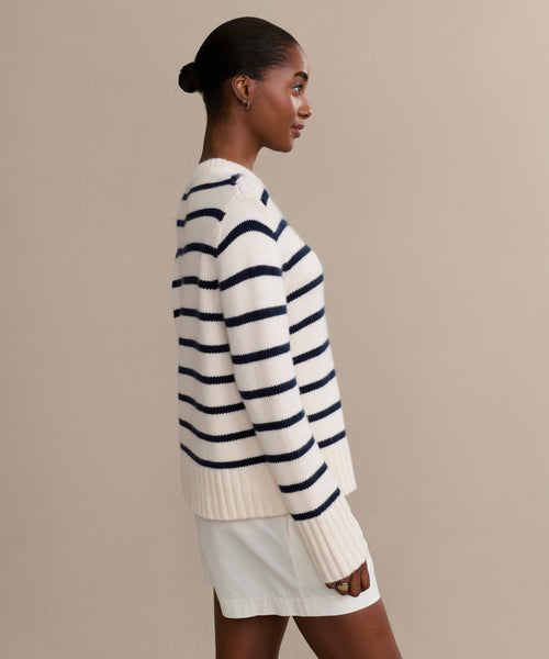 A woman with dark hair in a low bun models the Grace Crewneck, a luxury navy stripe knit sweater, paired with a white skirt as she stands in profile against a plain beige background.