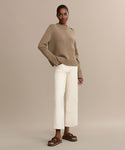 A woman stands against a beige background wearing the Grace Crewneck, a luxurious cashmere sweater with shoulder buttons, paired with off-white wide-leg pants and brown sandals. Her hair is pulled back, she smiles softly, and has one hand in her pocket.