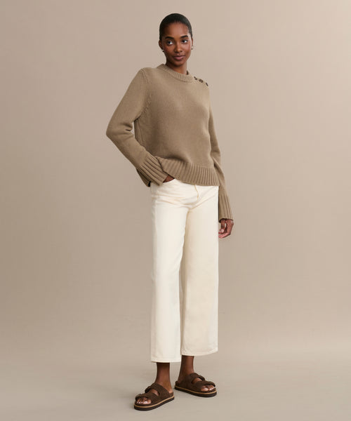 A woman stands against a beige background wearing the Grace Crewneck, a luxurious cashmere sweater with shoulder buttons, paired with off-white wide-leg pants and brown sandals. Her hair is pulled back, she smiles softly, and has one hand in her pocket.