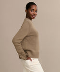 A woman with dark hair pulled back stands sideways with one hand in her pocket, wearing the luxurious Grace Crewneck tan cashmere sweater and white pants, set against a plain beige background.