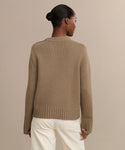 A woman with dark hair in a bun is seen from behind, wearing the Grace Crewneck—a luxurious taupe knit sweater with ribbed cuffs and hem—paired with off-white pants, standing against a plain beige background.