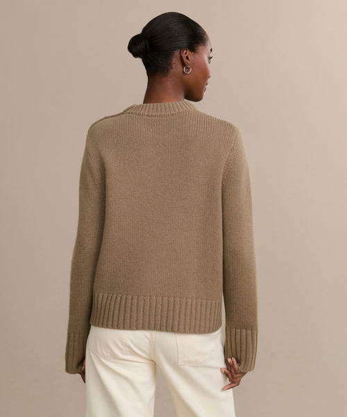 A woman with dark hair in a bun is seen from behind, wearing the Grace Crewneck—a luxurious taupe knit sweater with ribbed cuffs and hem—paired with off-white pants, standing against a plain beige background.