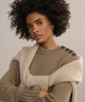 A woman with natural curly hair wears the luxurious Grace Crewneck knit tan sweater with large shoulder buttons, and a light beige sweater draped over her shoulders. She looks at the camera with a neutral expression against a plain background.