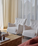 Two white, modern Greenwich Chairs with wooden legs in mini boucle fabric are placed side by side by a large window with sheer curtains. A beige coffee table with books and vases sits in front, and a peach sofa is partially visible.