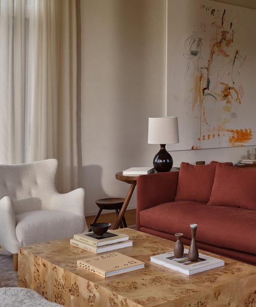 A contemporary living room with the Greenwich Chair in mini boucle, a rust sofa, a light wood coffee table with books and vases, a dark lamp on a wooden desk, sheer curtains, and a handmade abstract painting—made in the USA.
