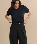 A woman with wavy brown hair wears a black t-shirt tucked into high-waisted, wide-leg Greta Trouser, standing against a beige background while adjusting her tailored trousers and looking downward.