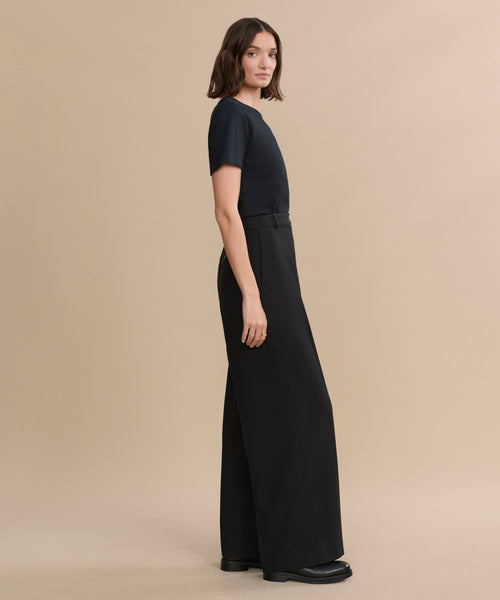 A woman stands sideways against a beige background, wearing a black short-sleeve top, the Greta Trouser—high-waisted, wide-leg tailored trousers in black with a relaxed drape—and black shoes.