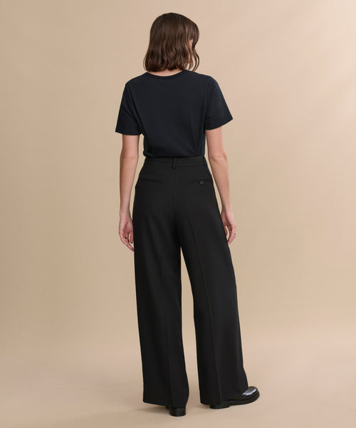 A person stands facing away, wearing a black t-shirt tucked into high-waisted Greta Trouser with a relaxed drape and black shoes, set against a plain beige background.