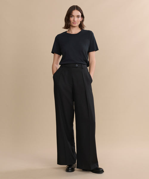 Against a plain beige background, a person wears a black t-shirt with Greta Trouser—tailored black trousers featuring a relaxed drape—paired with black shoes, hands in pockets and a neutral expression.