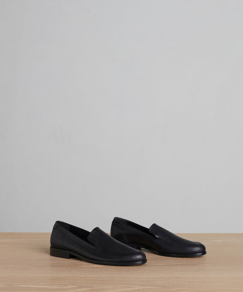 A pair of Griffin Loafer minimalist black shoes made from fine calf leather, displayed side by side on a light wood surface with a plain gray background.
