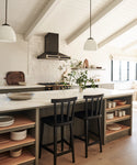 A modern kitchen features a white island with two Hampton Counter Stools in black oak, open shelves with dishes, pendant lights, a black range hood, and a branch in a vase. Sunlight from a large window fills the space with warmth.