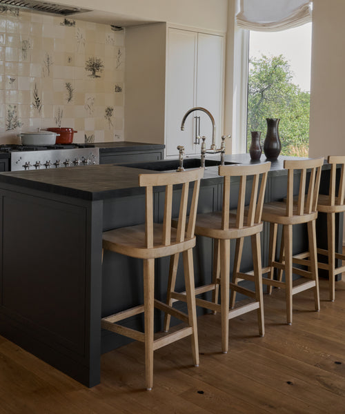 A modern kitchen features a dark island with a sink, Hampton Counter Stools along the counter, a light botanical-patterned tiled backsplash, and a large window framing views of lush greenery.