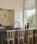 A modern kitchen showcases Hampton Counter Stools in light white oak at a dark island, double sink with gooseneck faucet, cream cabinets, botanical tile backsplash, and a large window with roman shade framing green trees.