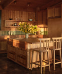 A sunlit wooden kitchen showcases a White Oak island paired with two Hampton Counter Stools, a green backsplash, and a vase of yellow flowers. Open shelves with bowls and cups enhance the warm natural wood cabinetry.